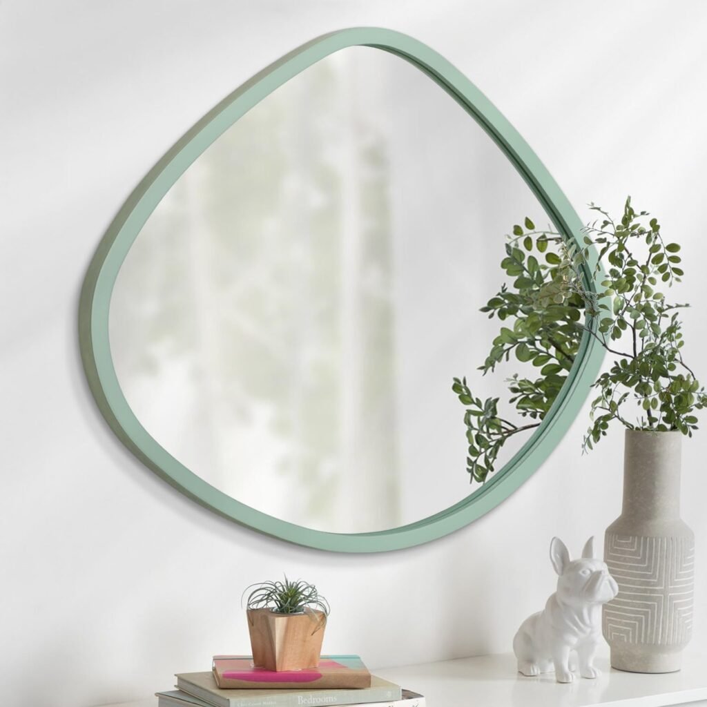 Soft-edged green-framed wall mirror above stacked books and vase with greenery