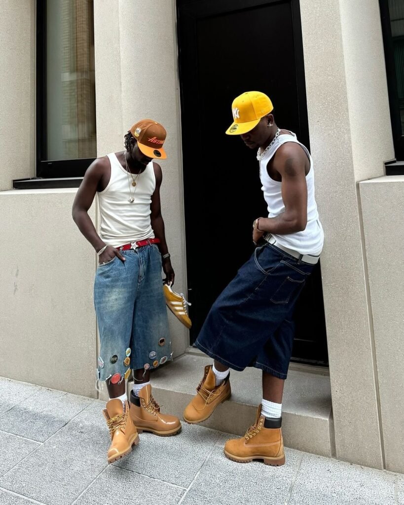 Two men in carpenter jorts, Timberland boots, and white tank tops walking on the street