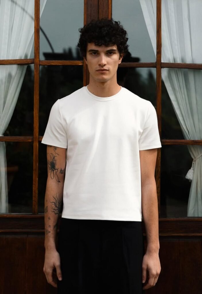 Model wearing structured white t-shirt and black trousers in front of window with sheer curtains