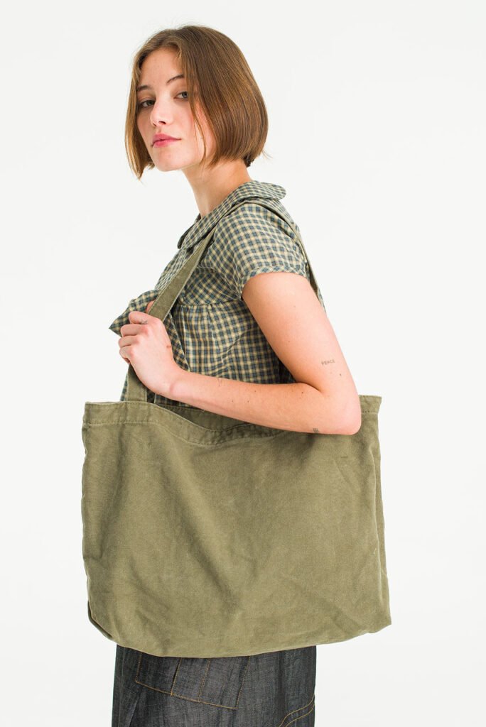 A young woman holding an oversized khaki canvas tote bag from Olive Clothing, wearing a gingham top.
