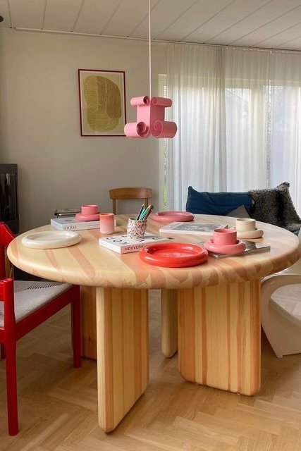 A pastel wood round table with sculptural curved legs, styled with pink tableware in a bright, modern interior — reminiscent of Gustav Westman’s furniture.