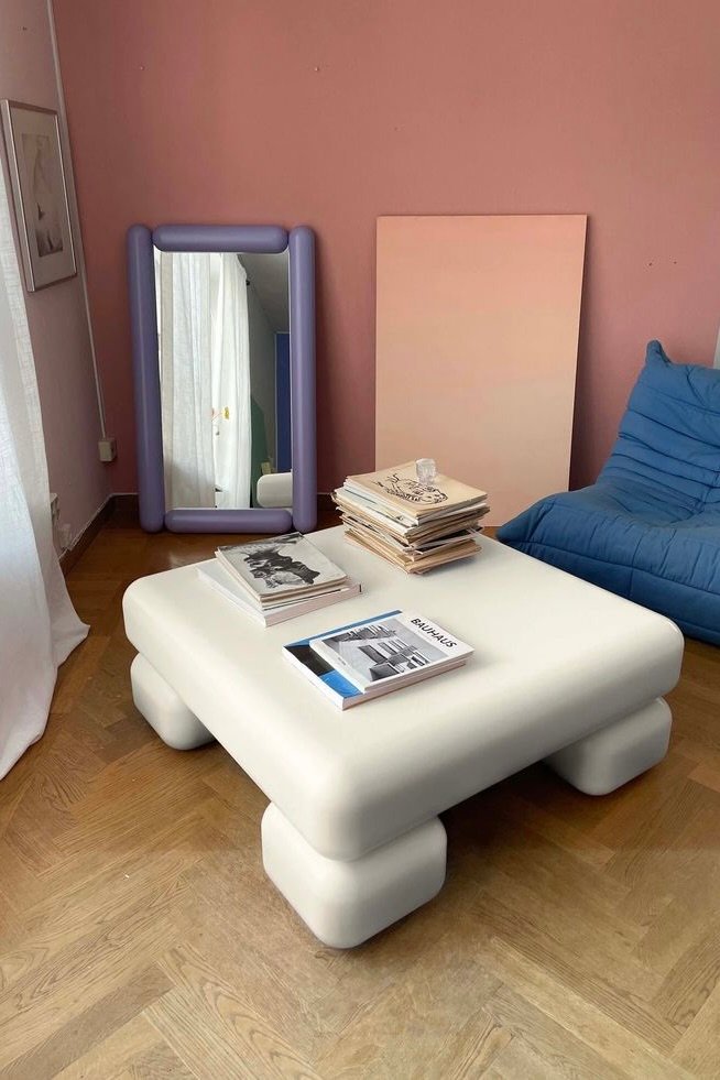 A soft white coffee table with rounded block legs, styled alongside a wavy purple mirror in a pastel pink interior — inspired by Gustav Westman’s design style.