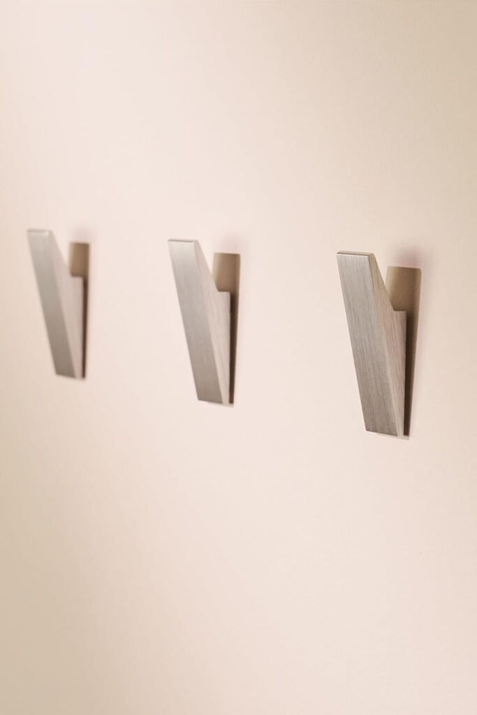 Four angled brushed silver wall hooks arranged evenly on a beige wall.